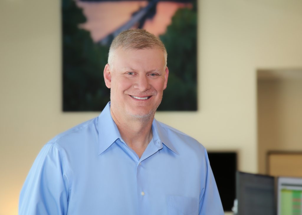 Man in a blue shirt smiling indoors with a painting in the background