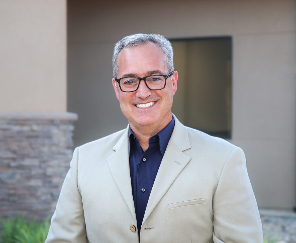 A man wearing a suit and glasses, smiling at the camera in a professional setting
