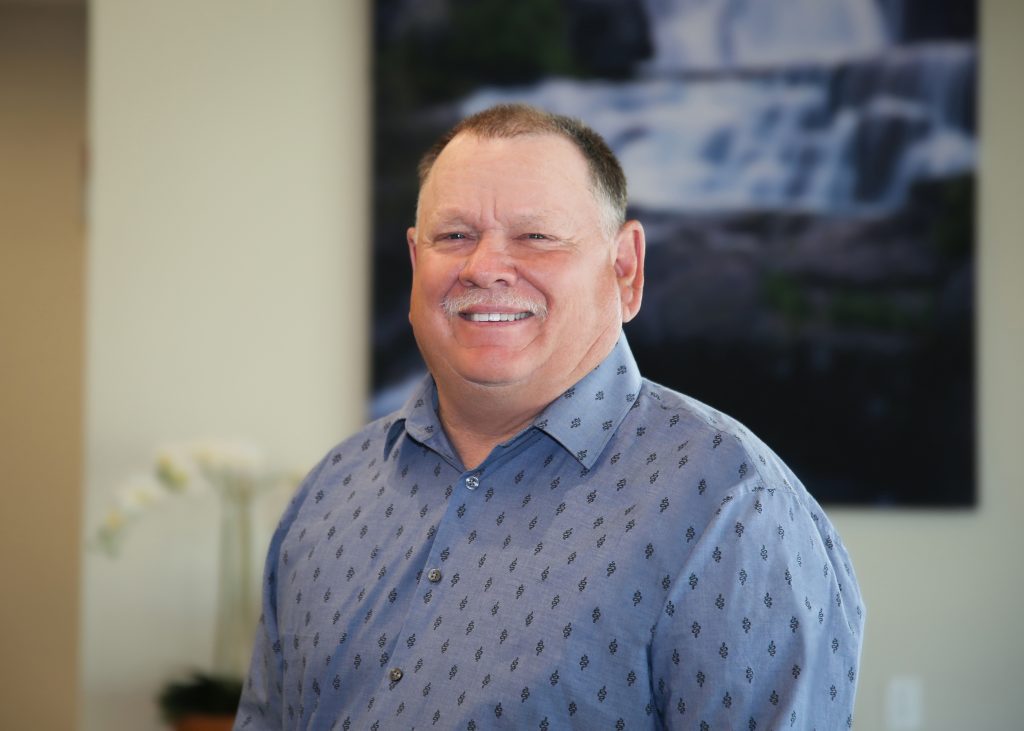 Man in a blue patterned shirt smiling indoors with a waterfall picture in the background