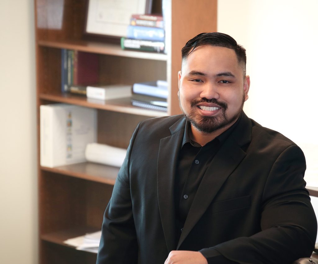 A man in a black suit smiling, with a background of office bookshelves