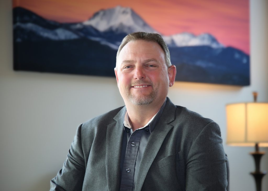 Man in a gray suit smiling indoors with a mountain painting in the background