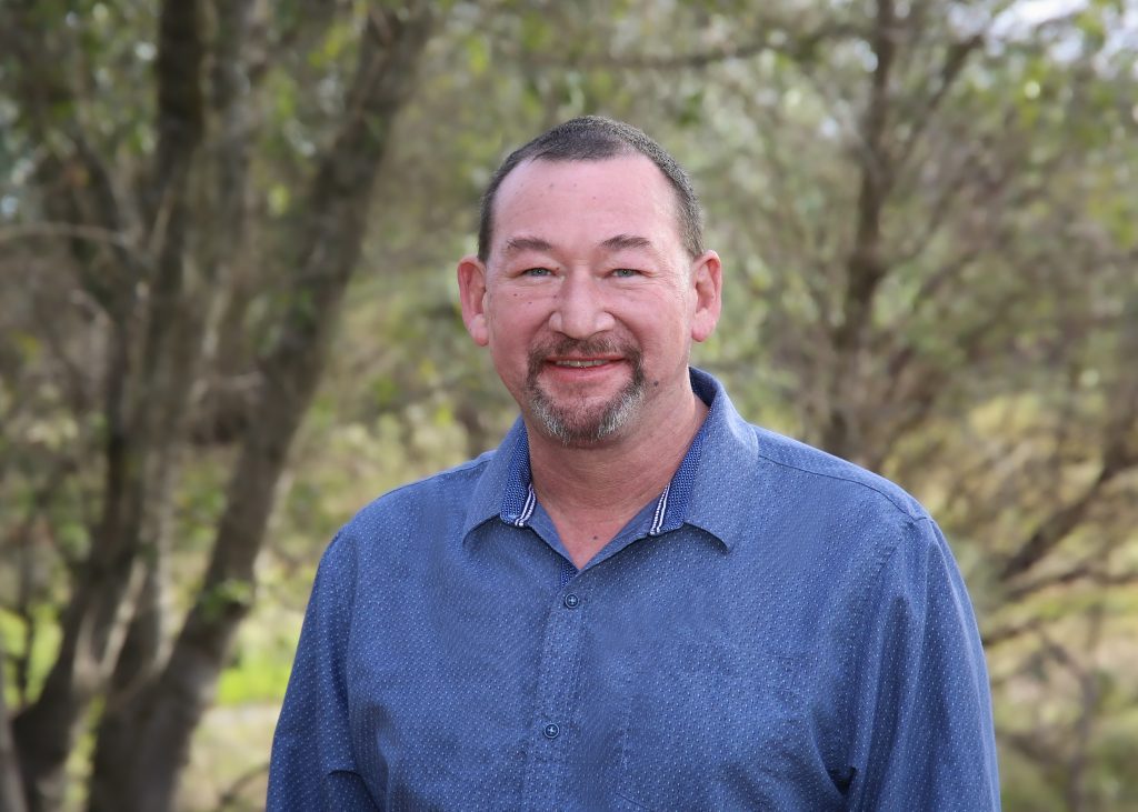 Man in a blue shirt smiling outdoors with trees in the background