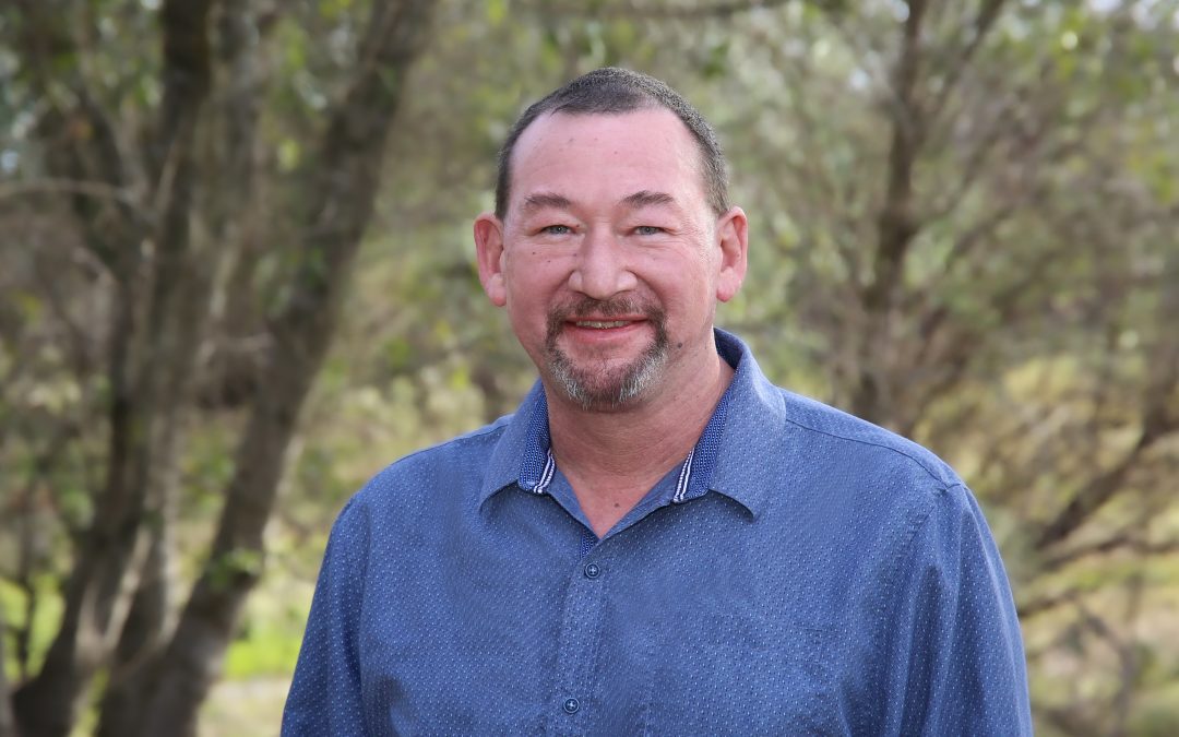 Man in a blue shirt smiling outdoors with trees in the background